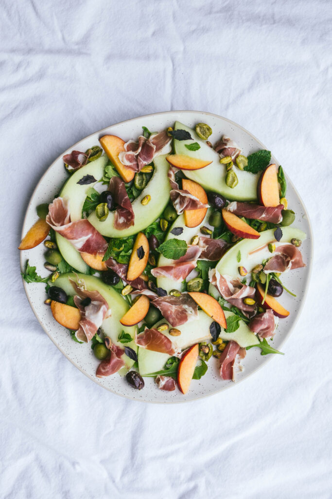 Prosciutto, melon, and peaches on a platter with olives and fresh herbs